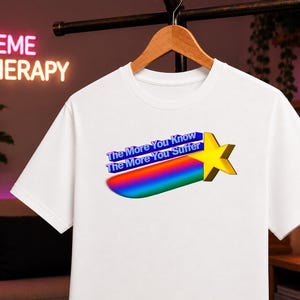 Può includere: Una t-shirt bianca a maniche corte con la grafica di una stella gialla con una scia color arcobaleno. Il testo "The More You Know" e "The More You Suffer" è stampato in blu sopra la scia arcobaleno in uno stile tridimensionale.