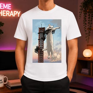 May include: A white t-shirt featuring a graphic print of a long, white cat standing upright on a tall, red metal industrial tower structure against a light blue sky with clouds. The cat is positioned vertically, gripping the tower structure.