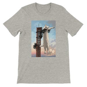 May include: A heather gray t-shirt featuring a graphic print of a giant white cat clinging to a tall, dark red industrial metal tower against a light blue and pink sky. The cat is positioned vertically, holding onto the structure with its front paws.