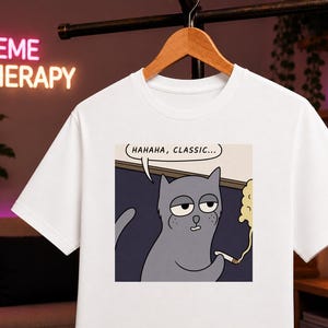 Op de afbeelding: Een wit t-shirt met korte mouwen en een grafische print van een grijze kat die een brandende sigaret vasthoudt. De kat kijkt verveeld en boven zijn hoofd staat een tekstballon met de tekst "HAHAHA, CLASSIC...". Het t-shirt hangt aan een houten kledinghanger.