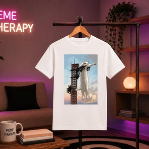 May include: A white short-sleeved t-shirt featuring a graphic print of a large white cat clinging to a tall, narrow, red-brown metal tower structure against a light blue sky with clouds. The shirt has a crew neckline and a standard straight hem.