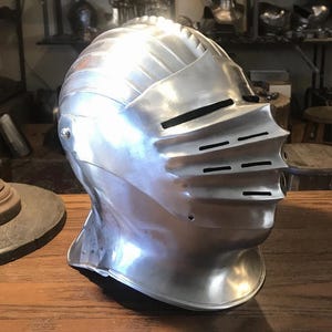 May include: A polished steel knight's helmet featuring a rounded dome, a hinged visor with horizontal eye and breathing slits, and a flared neck guard. The metallic surface reflects light, highlighting the curved contours of the protective headgear.