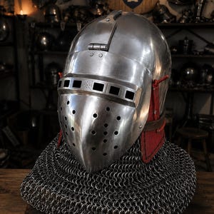 May include: A polished steel bascinet helmet featuring a hinged visor with rectangular eye slits and circular ventilation holes. The helmet is attached to a protective red leather neck guard and a heavy steel chainmail aventail covering the shoulders.