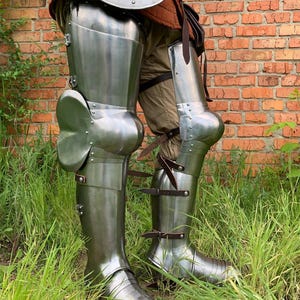 May include: Polished steel leg armor featuring articulated cuisses, poleyns with side wings, and greaves. The metal plates are secured with brown leather straps and buckles over tan fabric trousers, providing full coverage from the thigh to the foot.