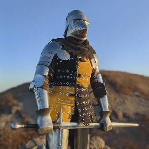 May include: A full suit of polished steel plate armour featuring a pointed visor helmet, chainmail neck protection, and a black and yellow quilted gambeson with metal studs. The armour includes articulated arm and leg pieces and a long steel sword.