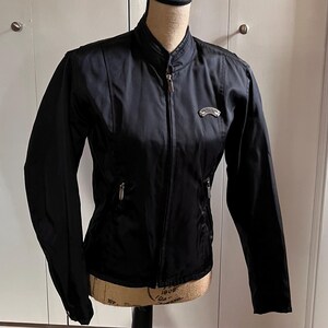 May include: Black motorcycle jacket with a stand-up collar and a silver zipper. The jacket has a small silver emblem on the chest and two zippered pockets. The jacket is displayed on a mannequin.