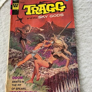 May include: A vintage comic book cover featuring a colorful illustration of a man and two women in a jungle setting. The title of the comic is "TRAGG and the SKY GODS" and the text below reads "DOOM AWAITS IN THE PIT OF SPEARS... "DEATH-DUEL!"  The comic is published by Western Publishing Company, Inc.