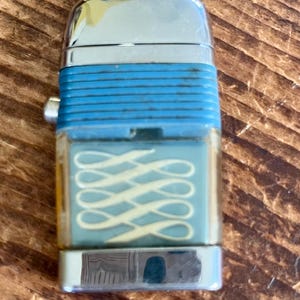 May include: A vintage lighter with a silver top and bottom, and a turquoise blue center section. The center section has a clear window with a repeating white and yellow design. The lighter is rectangular and sits on a wooden surface.