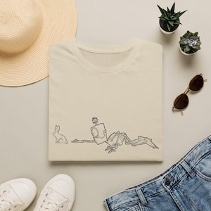 May include: A beige short-sleeved t-shirt featuring a black line art illustration of a robot lying on the ground facing a small rabbit. The robot has a single large circular eye and a small gear-like detail on top of its head.