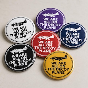 May include: A set of five circular pin-back buttons in white, purple, dark blue, red, and yellow. Each button features a graphic of a jet airplane and the text "WE ARE ALL ON THE DECOY PLANE" printed in a bold, sans-serif font with a circular border.