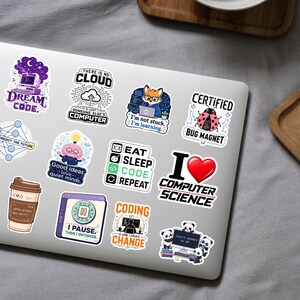 K&ouml;nnte beinhalten: Eine Sammlung von zw&ouml;lf bunten Vinyl-Aufklebern auf einem silbernen Laptop-Deckel. Die Designs zeigen verschiedene Illustrationen und Texte zum Thema Programmierung, darunter Schriftz&uuml;ge wie Dream in Code, Eat Sleep Code Repeat und I Love Computer Science.