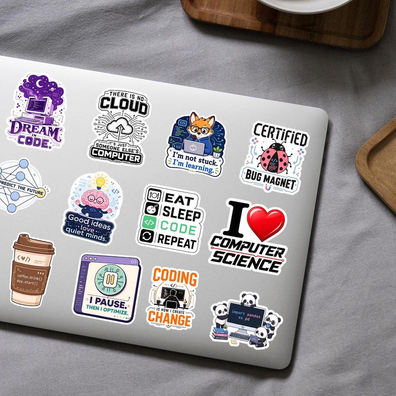 K&ouml;nnte beinhalten: Eine Sammlung von zw&ouml;lf bunten Vinyl-Aufklebern auf einem silbernen Laptop-Deckel. Die Designs zeigen verschiedene Illustrationen und Texte zum Thema Programmierung, darunter Schriftz&uuml;ge wie Dream in Code, Eat Sleep Code Repeat und I Love Computer Science.