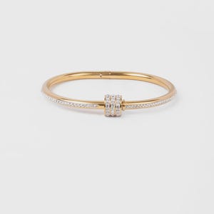 May include: A gold-toned hinged bangle bracelet featuring a central cylindrical charm adorned with three rows of clear crystals. The outer surface of the bangle is also inlaid with a continuous line of small, sparkling clear crystals.
