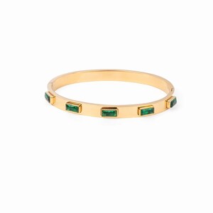 May include: A gold-toned hinged bangle bracelet featuring five rectangular, emerald-green stones set at intervals along the outer surface. The bracelet has a smooth, polished finish and a secure clasp mechanism visible on the side of the band.