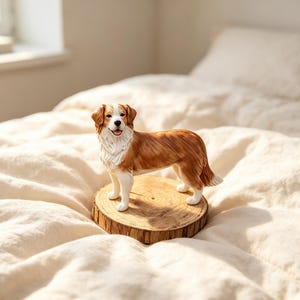 May include: A standing dog figurine with a brown and white coat, featuring a detailed textured fur pattern, a white chest, and a black nose. The dog is positioned on a circular, light-coloured wooden slice base with visible natural wood grain rings.