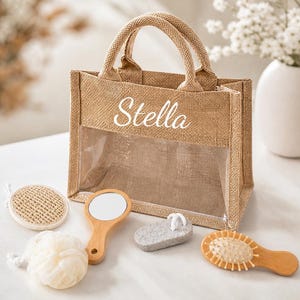 May include: A tan burlap tote bag with the name Stella printed in white script on the front, featuring a clear plastic window. The set includes a round jute scrub pad, a white mesh bath sponge, a wooden hand mirror, a gray pumice stone, and a brush.
