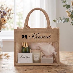 May include: A tan burlap tote bag with two handles, featuring a black bow icon and the name Krystal printed on the front. Inside the bag is a bottle, a light-colored sleep mask, a scented candle, a small ring box, and a Thank You card.