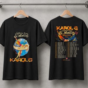 以下が含まれることがあります： 前面にカラフルな鳥の羽と「Viajando Por El Mundo」「Karol G」の文字が描かれた地球儀のグラフィックが入った黒いTシャツ。背面には「Karol G」のロゴ、「Tropitour 2026」の文字、ツアーの日程と場所のリストがプリントされています。