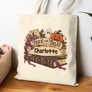 Op de afbeelding: Een crèmekleurige canvas tas met een bedrukte afbeelding van een boek, een pompoen, een spook en bloemen. Een banner in het midden toont de tekst "TRICK OR TREAT" boven de naam "Charlotte" in een donker, gestileerd lettertype.