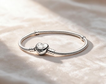 May include: A silver snake chain bracelet featuring a heart-shaped clasp. The clasp is engraved with the text PANDORA. The bracelet has two fixed stations along the chain, designed to hold charms in place, and a polished metallic finish throughout.