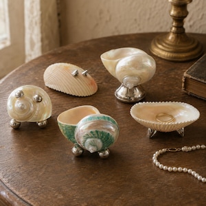 May include: A collection of decorative seashell containers and dishes featuring iridescent surfaces, some with green accents, supported by small polished silver-toned metal ball feet or pedestals. One shell dish contains a simple gold-toned ring.