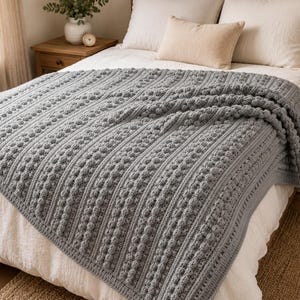 Pode incluir: Uma manta de crochê cinza com um padrão repetitivo de colunas verticais de pontos pipoca texturizados e seções caneladas lisas. A manta está sobre uma cama com roupa de cama branca, destacando o trabalho detalhado do fio e a cor cinza.