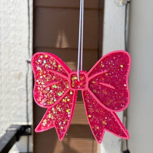 May include: A bright pink bow-shaped ornament featuring a textured surface embedded with multi-colored iridescent glitter. The bow is suspended by a thin white elastic cord attached to the center, creating a decorative hanging piece.
