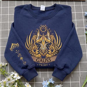 May include: A navy blue crewneck sweatshirt featuring a large, intricate gold and white embroidered graphic on the chest. The design depicts a stylised mechanical helmet with wings and the text "GAUSS PRIME". A matching gold symbol is on the left sleeve.