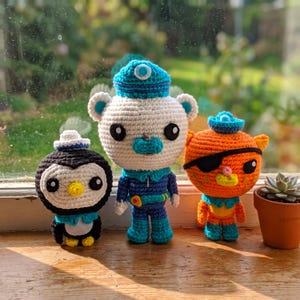 May include: Three handmade crochet dolls featuring a black and white penguin with a blue hat, a white polar bear in a blue uniform with a matching cap, and an orange cat with a black eye patch and blue hat, standing in a row.