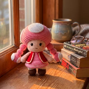 Puede incluir: Una muñeca de ganchillo con un gran sombrero rosa en forma de seta con un círculo blanco en la parte superior, dos trenzas rosas, un chaleco rojo sobre un vestido rosa y zapatos marrones. La muñeca tiene ojos negros y una boca pequeña.