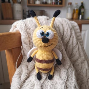 May include: A handmade crochet bee toy featuring a round yellow head with large white eyes, blue pupils, and black antennae. The body has yellow and brown horizontal stripes, small white wings, and black extremities. The toy has a stitched smile.
