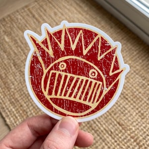 May include: A circular sticker featuring a stylized red face with a distressed, weathered texture. The design includes a large, wide-grinning mouth with vertical teeth, two small circular eyes, and a spiky, flame-like crown surrounding the head.