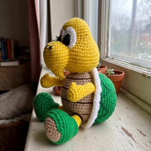 May include: A crocheted turtle figure featuring a yellow head with white and black eyes, a tan belly with brown horizontal lines, green feet, and a green shell with a white border. The figure is crafted with visible yarn stitches throughout.