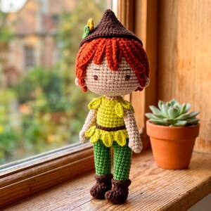 Puede incluir: Una muñeca de ganchillo hecha a mano con un sombrero puntiagudo marrón con un pequeño detalle de hoja amarilla y verde, pelo naranja brillante, cara beige claro, top amarillo con dobladillo en forma de hoja, pantalones verdes y botas marrones.