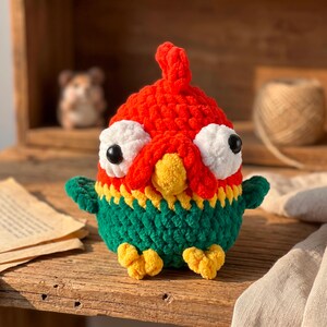 May include: A crochet plush toy shaped like a bird with a bright red head, a yellow beak, and a green body. The bird features large white eyes with black pupils, small green wings on the sides, and two yellow feet at the bottom of the round body.