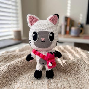 Könnte beinhalten: Ein weiß-schwarzes gehäkeltes Katzen-Stofftier mit rosa Innenohren und einer kleinen rosa herzförmigen Nase. Die Katze hat große schwarze Augen und trägt eine leuchtend rosa gehäkelte Umhängetasche mit einem kleinen weißen Detail an der Vorderklappe.