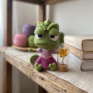 May include: A green crocheted chameleon doll with large eyes and a textured head, wearing a purple and pink dress with a lace-up bodice. Beside the doll is a small yellow crocheted flower inside a clear glass cloche with a cork base.