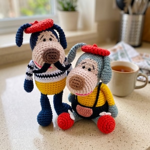 May include: Two handmade crochet dog dolls. The standing dog has dark blue ears, a red beret, a black moustache, and a white shirt with blue stripes over yellow trousers. The sitting dog has light blue ears, a red beret, yellow shirt, and black overalls.