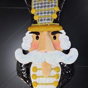 May include: A decorative nutcracker wall hanging featuring a black sequined hat and jacket, a white glittery mustache and hair, and gold trim accents. The figure has a tan face, black eyes, and a white vest with three gold circular buttons.
