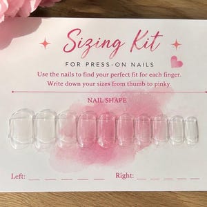 May include: A white rectangular sizing kit card for press-on nails with pink text and watercolor accents. The card features the title Sizing Kit, instructions, and ten clear, transparent plastic nail tips arranged in a horizontal row.