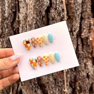 May include: Two sets of five almond-shaped press-on nails displayed on a white card. Each set features a variety of designs including a brown and orange checkered pattern, a brown nail with white flowers, an orange floral print, a cream swirl, and blue with white dots.