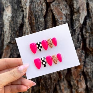 May include: A set of ten almond-shaped press-on nails displayed on a white card. The nails feature a glossy finish with a mix of solid hot pink, black and white checkered patterns, and tan and black leopard print designs arranged in two rows.