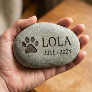 May include: A smooth, oval-shaped gray river stone featuring an engraved design. The stone displays a paw print icon on the left side, followed by the name LOLA and the years 2011 - 2024 engraved in a clear, dark, serif font on the surface.