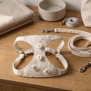 May include: A cream-colored pet harness and matching leash set featuring a textured fabric with embroidered daisy patterns and three-dimensional daisy appliqués. The harness includes adjustable metal buckles and a silver-toned metal leash clip.