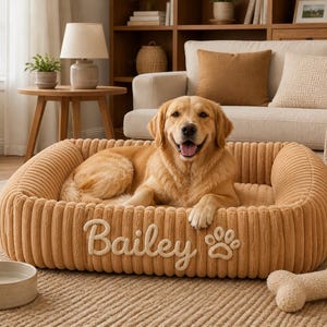 May include: A golden-coloured, rectangular pet bed with a ribbed, corduroy-like texture. The front of the bed features the embroidered name "Bailey" in a cursive script next to a paw print icon. A golden retriever is resting inside the bed.