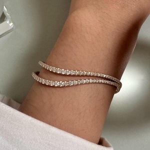 May include: A rose gold wavy bracelet featuring a continuous row of small, round, clear sparkling stones set along the entire length of the band. The design follows a gentle, undulating curve that wraps around the wrist, showcasing a polished metallic finish and a uniform arrangement of stones that catch the light.