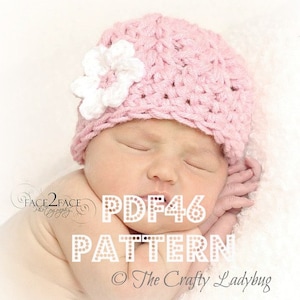 Può includere: Un berretto per neonato in crochet rosa con un fiore in crochet bianco sulla cima. Il berretto è sulla testa di un neonato. Il testo "PDF46 PATTERN © The Crafty Ladybug" è sull'immagine.