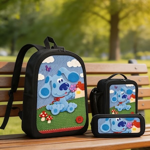 May include: A black backpack, lunch bag, and pencil case set featuring a blue dog character on a green grassy field with red mushrooms, white clouds, and a small house. The design includes a red button accent and a purple butterfly graphic.