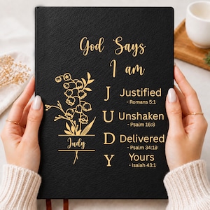 Op de afbeelding: Een zwart dagboek met gouden tekst en een bloemenillustratie. De kaft bevat de tekst God Says I am, gevolgd door de naam Judy verticaal, waarbij elke letter bij een woord hoort: J Justified, U Unshaken, D Delivered, Y Yours, en bijbelverzen.