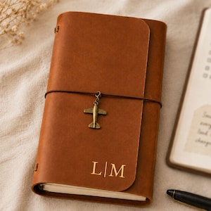 Puede incluir: Una funda de diario de cuero color canela con un monograma dorado en relieve con las letras L y M. Un cordón elástico marrón oscuro envuelve el centro, sujetando un pequeño dije de avión color latón antiguo. Tiene un acabado mate suave.